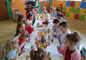 Dzieci z grupy Wiewiórek siedzą przy wigilijnym stole nakrytym białym obrusem na którym znajdują się świąteczne dekoracje, owocowo-słodki poczęstunek. W tle widać kolorową choinkę i zimowo-świąteczne dekoracje.